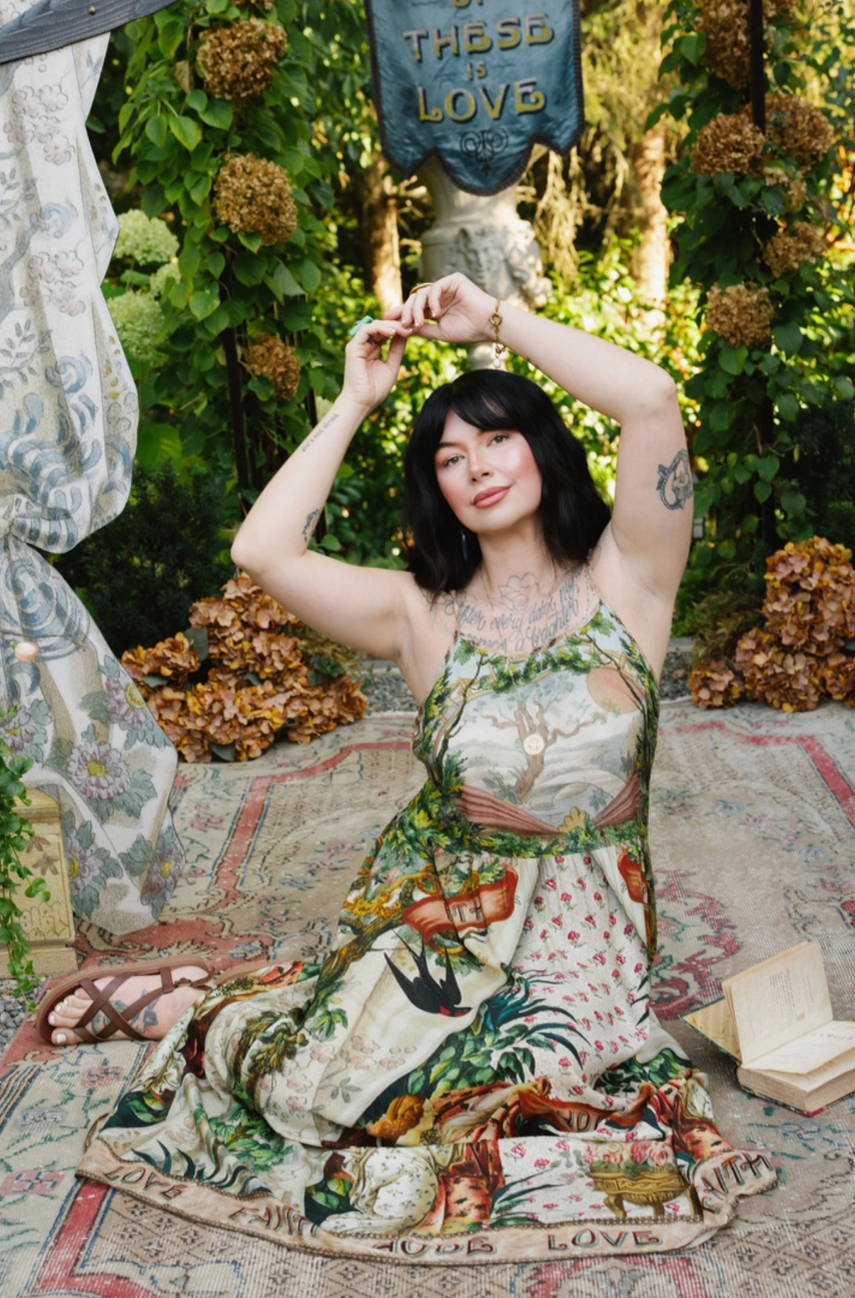  Model seated with arms raised in the Book of Love maxi dress, its illustrated skirt spread beside an open book | Bliss by Cori
