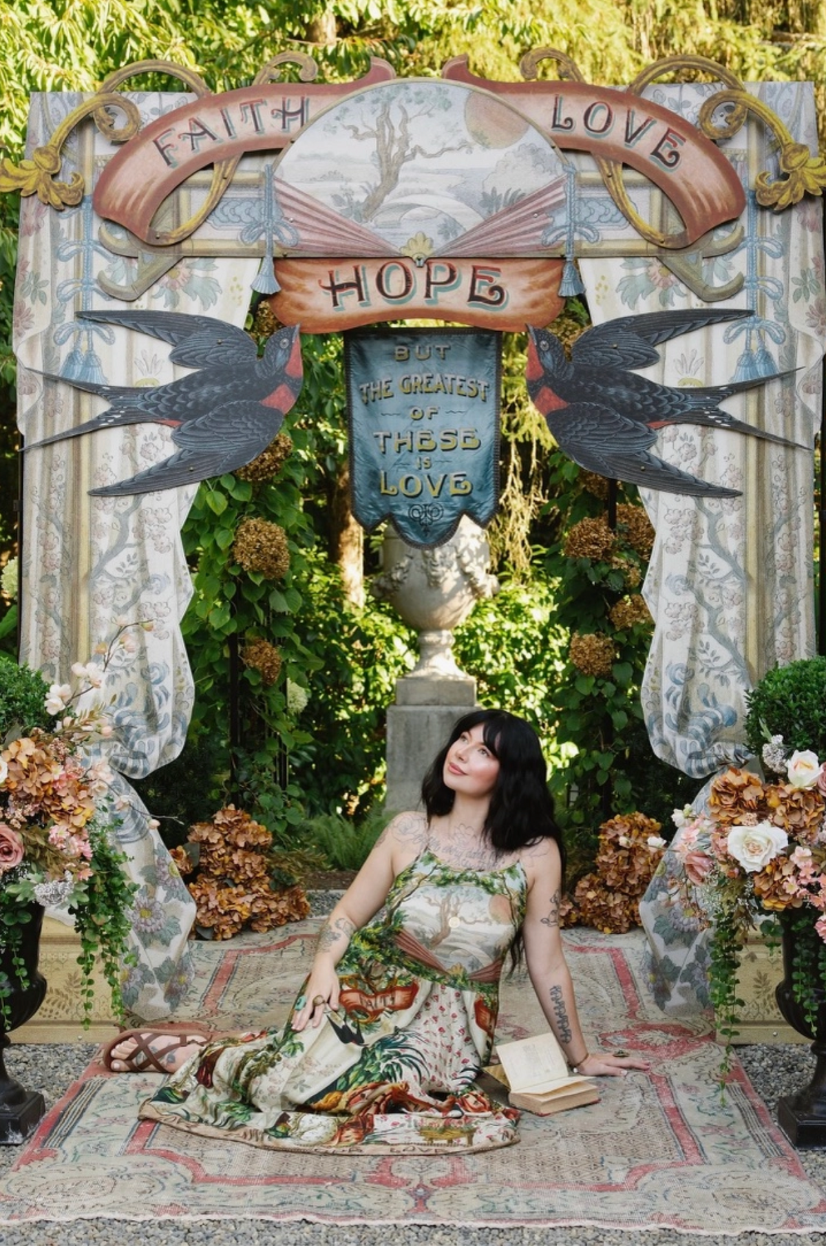 Model seated beneath a decorative garden arch in the Book of Love maxi dress, with skirt spread on a rug | Bliss by Cori