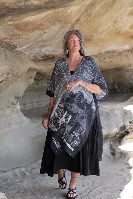 Woman walking through sandstone cave wearing Stargazer Goddess Duster Kimono with Leonid meteor shower and village scene print over black dress | Bliss by Cori Boutique