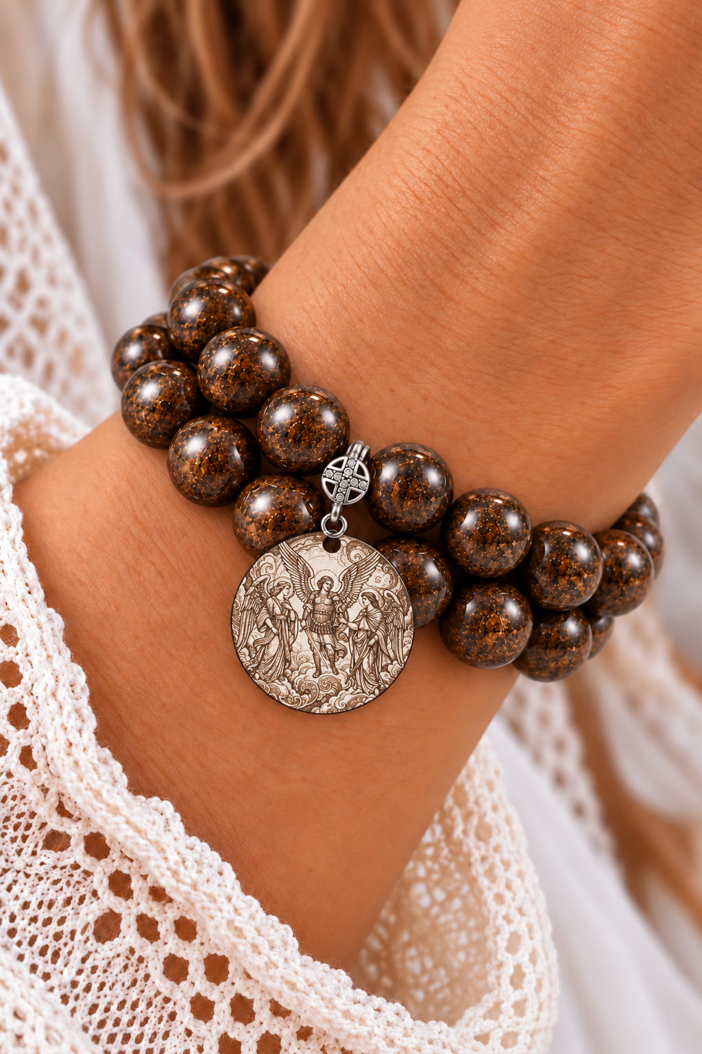 Two stacked brown bronzite bead bracelets with a round medal depicting Archangels Michael, Raphael, and Gabriel, worn beside a white lace sleeve | Bliss by Cori