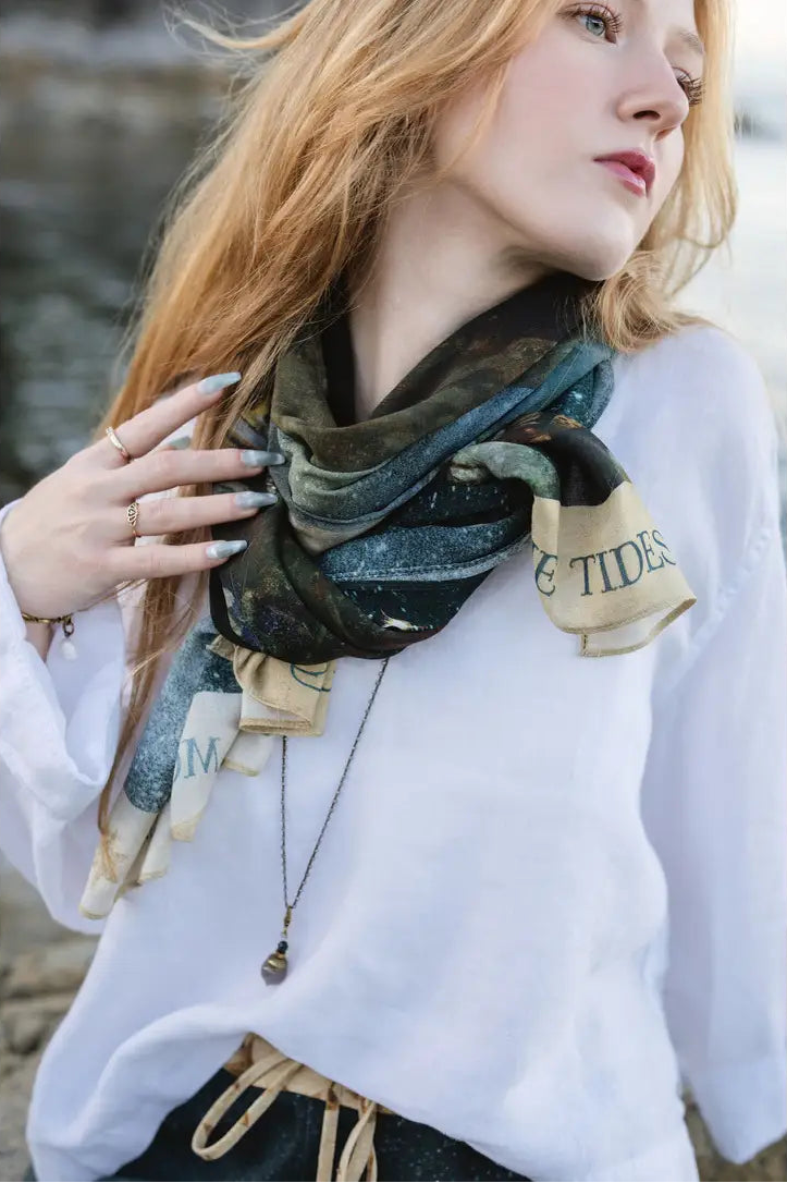 A woman with long red hair models a patterned scarf with 'E TIDES' printed on a beige tab, paired with a white top and black skirt in an outdoor setting | Bliss by Cori