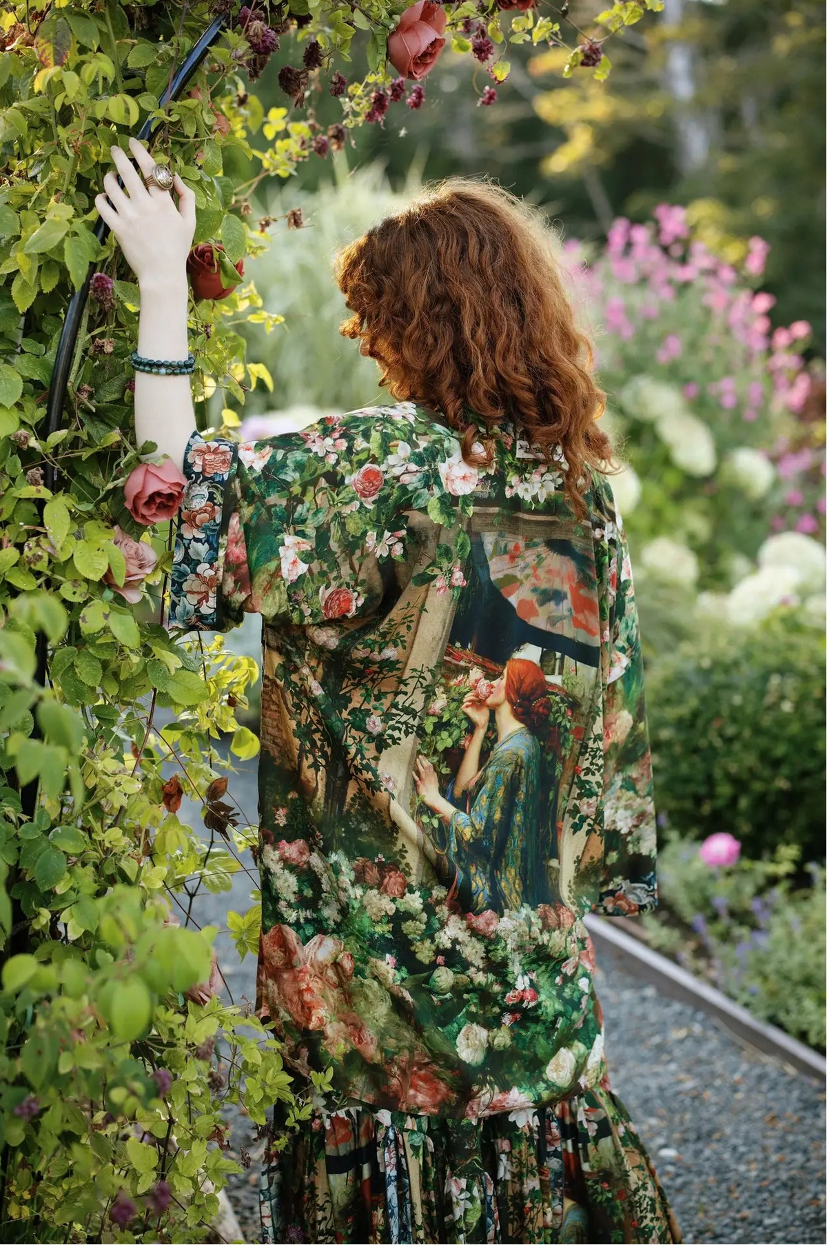 A woman with curly red hair stands in a garden, viewed from behind, wearing a short-sleeve button-up shirt and matching skirt with a detailed vintage-style floral and portrait print. The print features green foliage, pink and red flowers, and a painting of a woman with red hair | Bliss by Cori