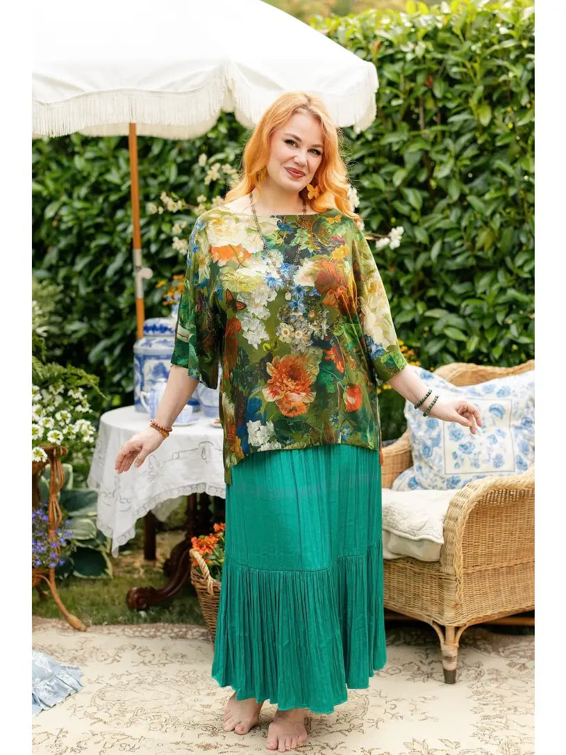 Red-haired woman wearing a green floral-and-bird print Luxe Tee tunic over a teal pleated skirt in a flower-filled garden | Bliss by Cori