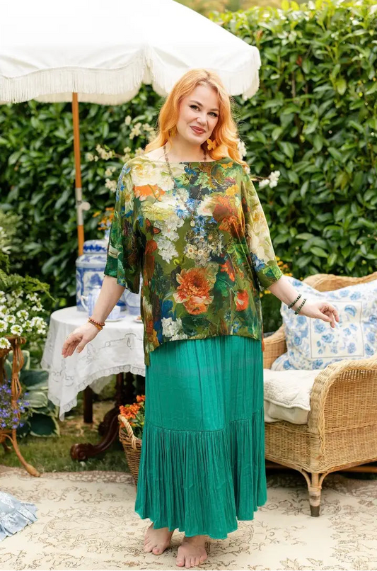 Red-haired woman wearing a green floral-and-bird print Luxe Tee tunic over a teal pleated skirt in a flower-filled garden | Bliss by Cori