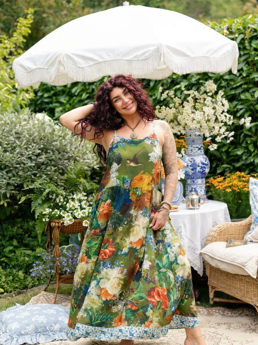 A woman wearing a green floral maxi dress stands outdoors under a white parasol in a garden setting with a table, chairs, and blue-and-white ceramic vase in the background | Bliss by Cori