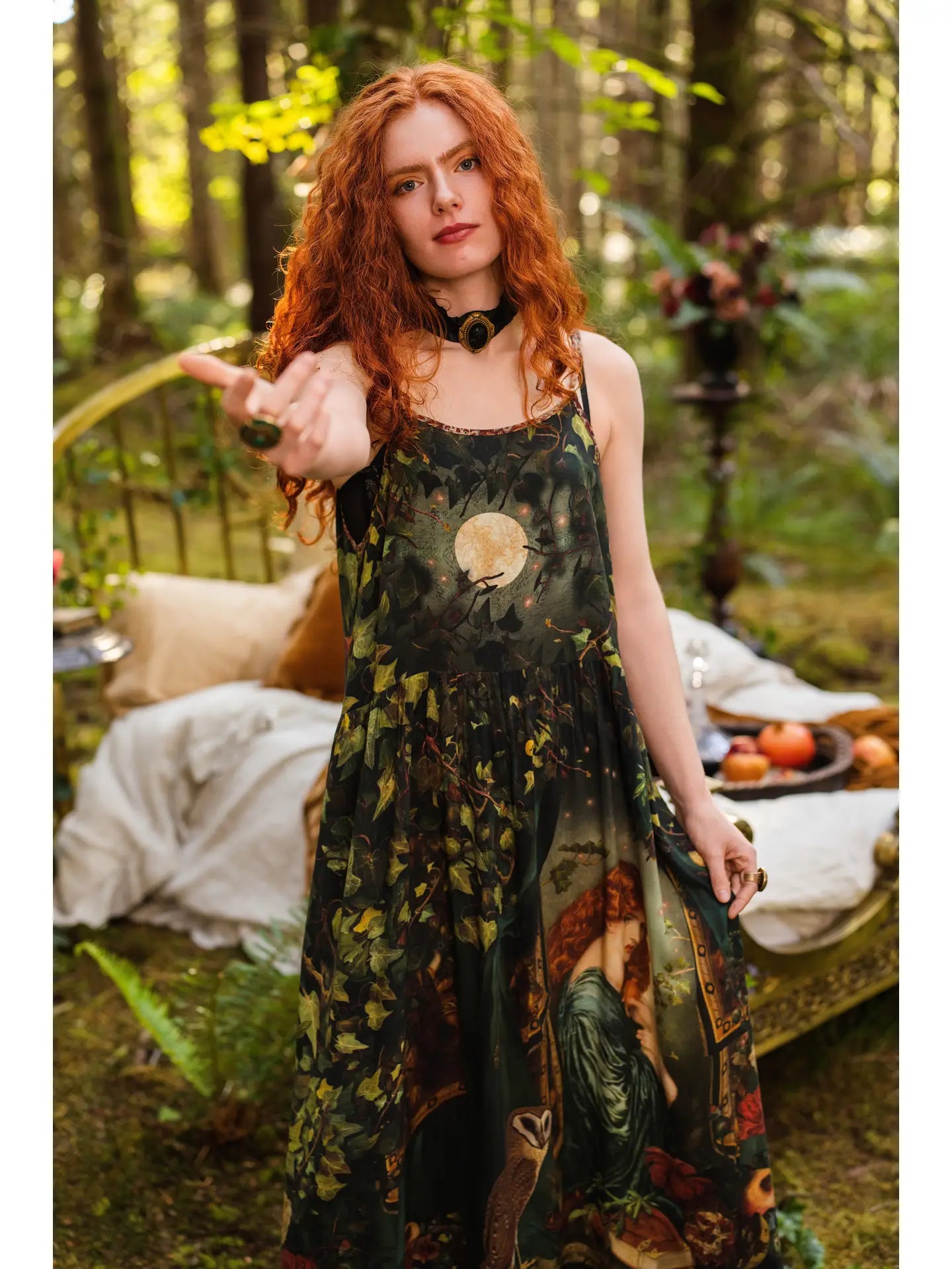 Front view of red-haired model in enchanted forest holding a pomegranate wearing Solstice Bohéme Maxi Slip Dress, moon and ivy print on bodice, Proserpine portrait on skirt, barn owl visible in background — Bliss by Cori