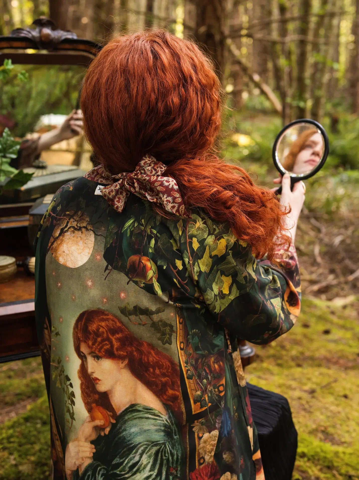 Back view of red-haired model in mossy forest holding hand mirror wearing Solstice Matinée Mid-Length Duster Kimono, full Rossetti Proserpine and moon print on back, antique dresser and ferns behind — Bliss by Cori