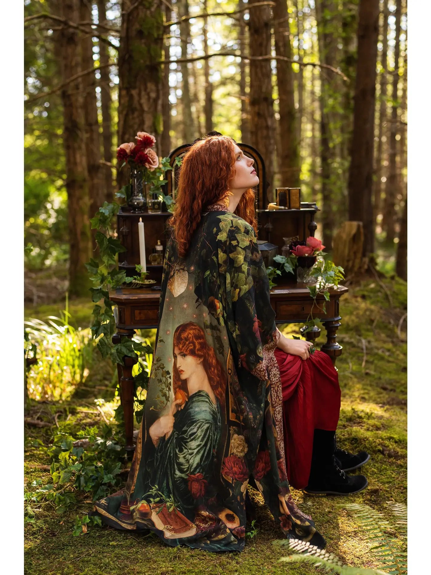 Model seated in forest wearing Solstice Opera Duster Kimono draped open, Rossetti Proserpine print cascading to the ground, antique writing desk with candles and roses in background — Bliss by Cori
