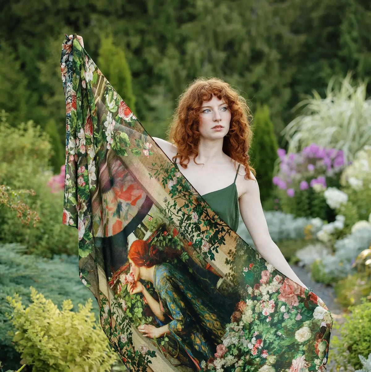 A woman with red hair stands in a lush garden holding up a large sheer scarf printed with a vintage-style floral and figure illustration. The scarf features green foliage, pink and red flowers, and a woman in a blue dress, layered over a dark green spaghetti-strap dress | Bliss by Cori