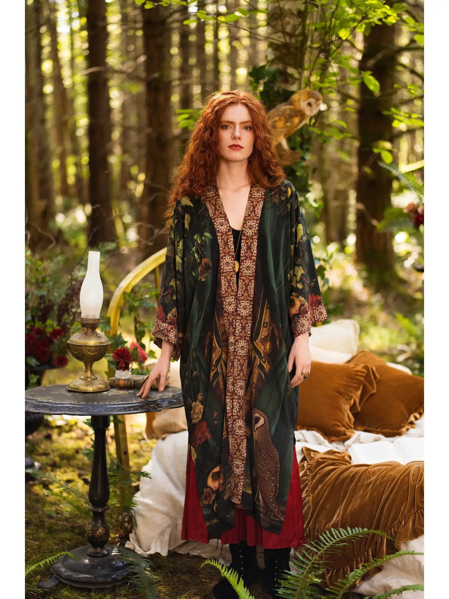 Front view of red-haired model in enchanted forest setting wearing Solstice Opera Duster Kimono open over burgundy dress, owl perched in tree canopy, oil lamp and velvet cushions nearby — Bliss by Cori