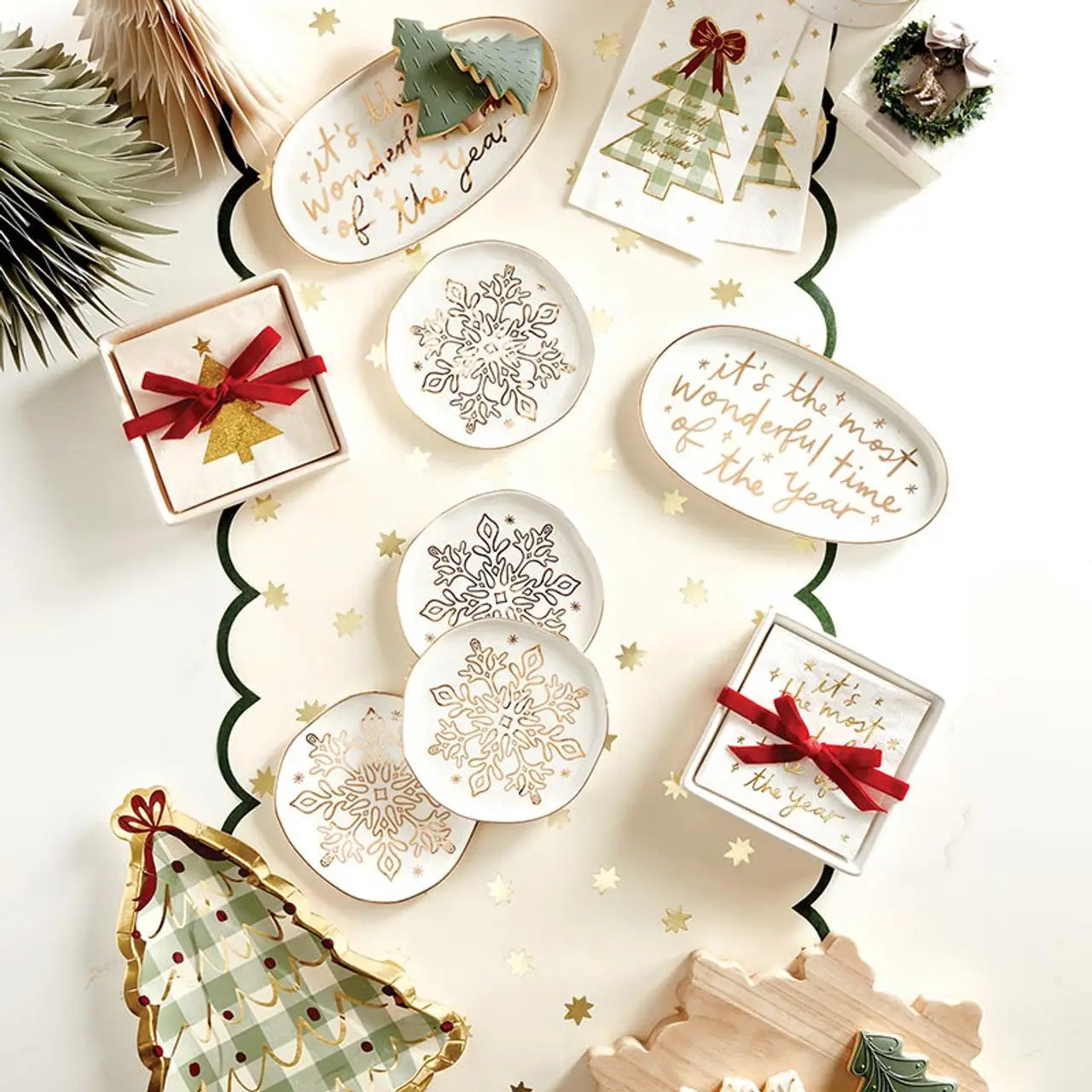 Overhead flat lay of holiday ceramic platters and coasters with gold snowflake designs and 'It's the Most Wonderful Time of the Year' gold foil text, styled with Christmas trees, gift boxes and star confetti – Bliss by Cori
