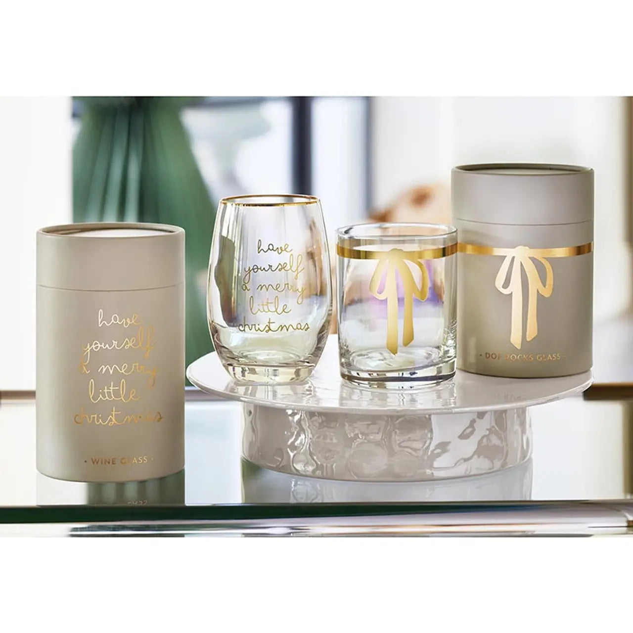 Merry Little Christmas iridescent stemless wine glass and rocks glass with gold script displayed on a marble tray beside matching taupe gift tube packaging – Bliss by Cori
