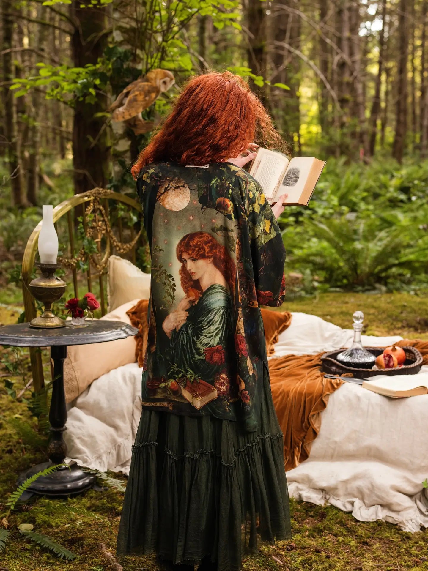 Back view of red-haired model in sunlit forest reading a book wearing Solstice Matinée Mid-Length Duster Kimono, Rossetti Proserpine portrait print visible, barn owl perched in canopy, oil lamp and pomegranate on bedside table — Bliss by Cori