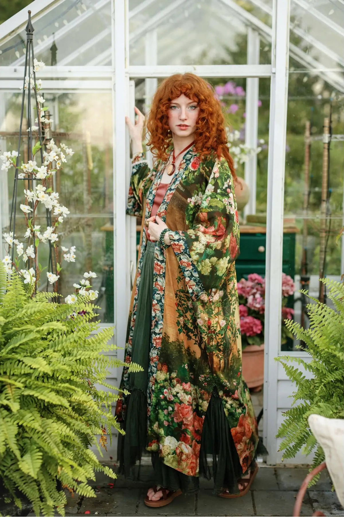 A woman with vibrant red hair stands in a greenhouse wearing a long-sleeved, bohemian-style floral kimono robe over a green dress. The robe features a dense multicolor floral pattern with green, orange, red, and yellow tones | Bliss by Cori