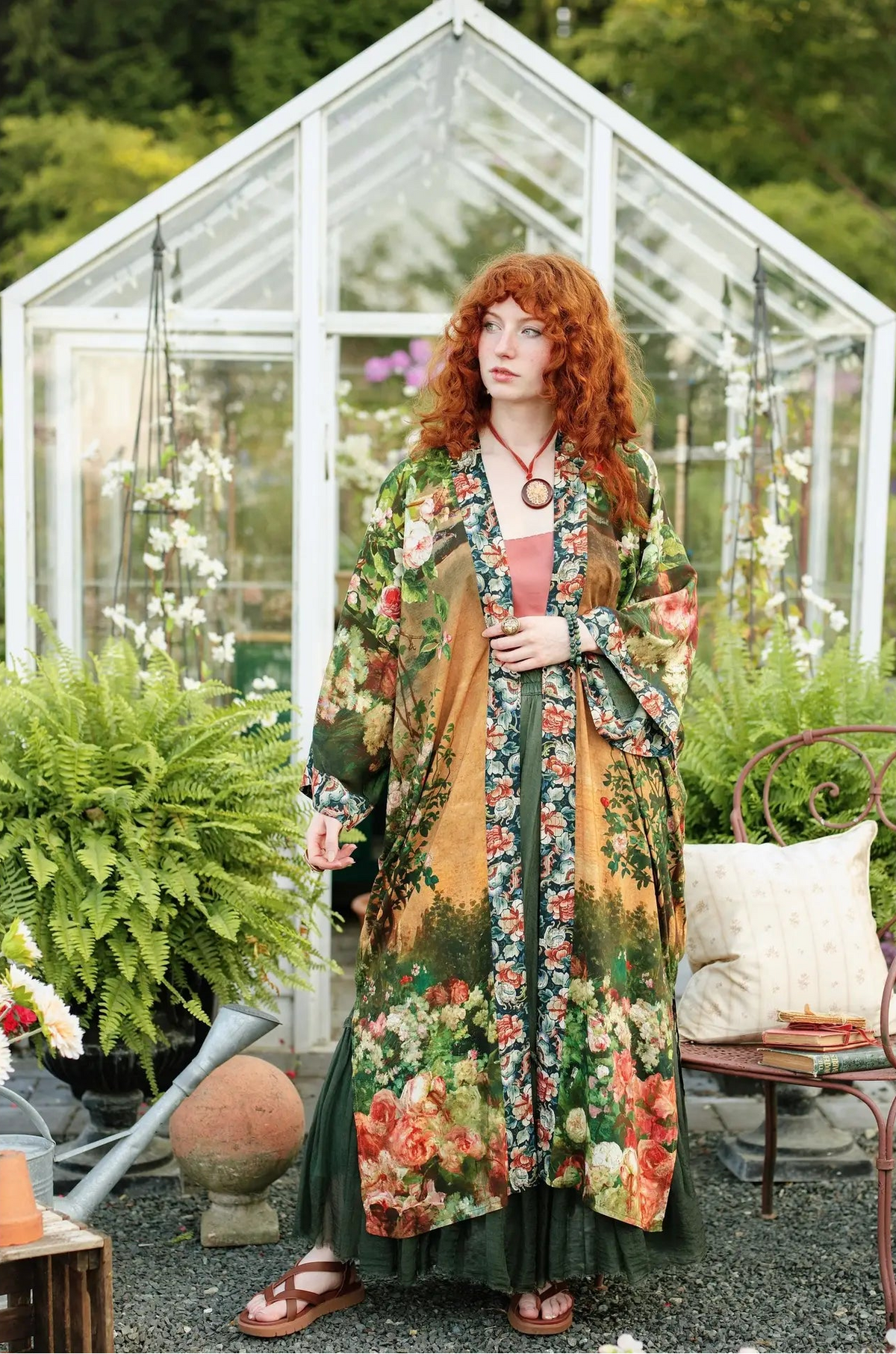 A woman with red hair stands in a greenhouse garden wearing a long, flowing floral robe over a pink top and dark green skirt. The robe features a warm mustard-yellow base with dense red, orange, and green botanical patterns | Bliss by Cori