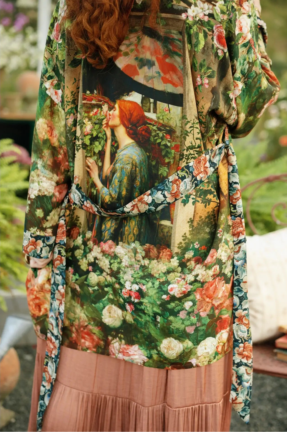 A close-up of a person wearing a long-sleeve floral kimono-style top with a large vintage-style art print of a woman in a garden, paired with a pleated dusty pink skirt in an outdoor garden setting | Bliss by Cori