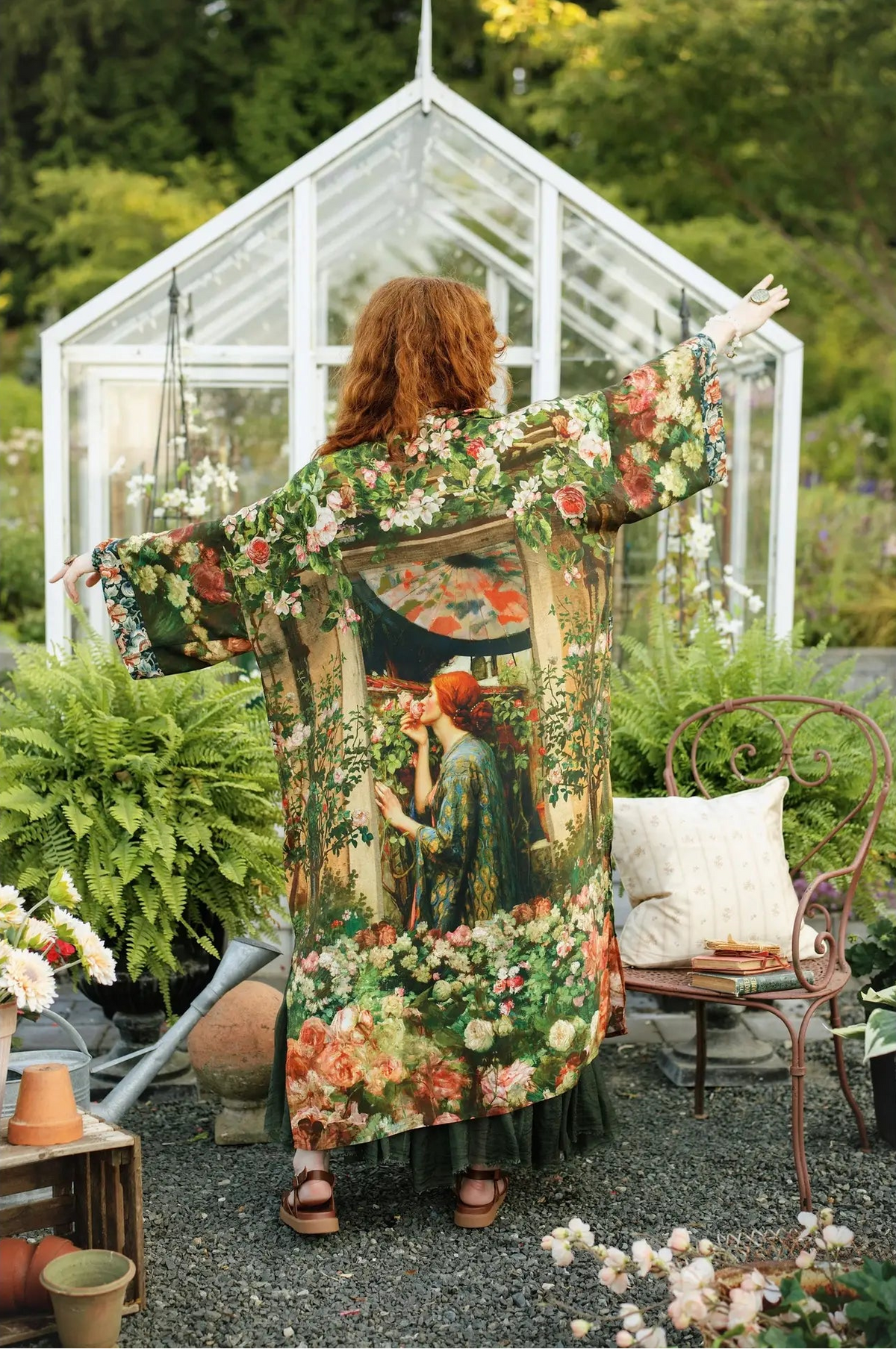 A woman stands with her back to the camera in a garden greenhouse setting, wearing a long-sleeved, wide-leg kimono-style robe covered in a dense floral and botanical print with an illustration of a woman in a green dress | Bliss by Cori