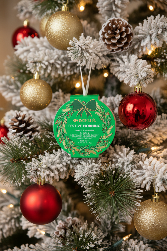 Spongellé Festive Morning Sweet Ambrosia body buffer ornament hanging on a snow-flocked Christmas tree with warm golden bokeh lights — Bliss by Cori Boutique