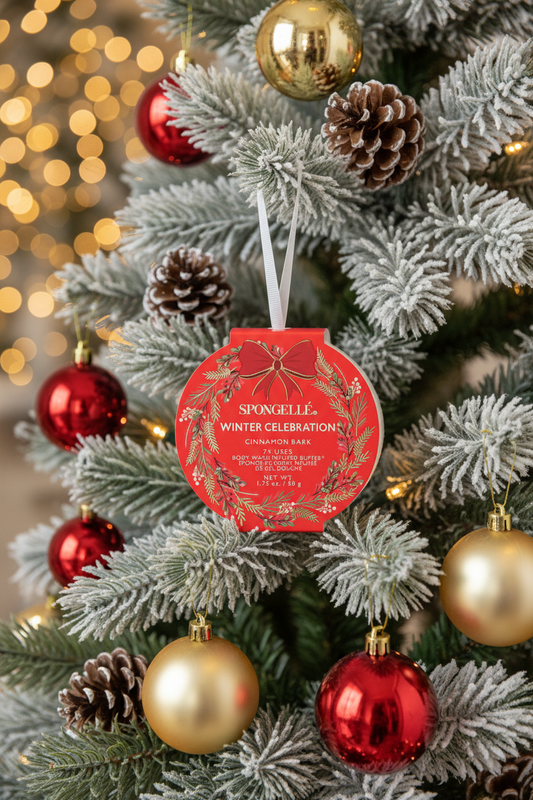 Spongellé Winter Celebration Cinnamon Bark body buffer ornament hanging on a snow-flocked Christmas tree with warm golden bokeh lights — Bliss by Cori Boutique